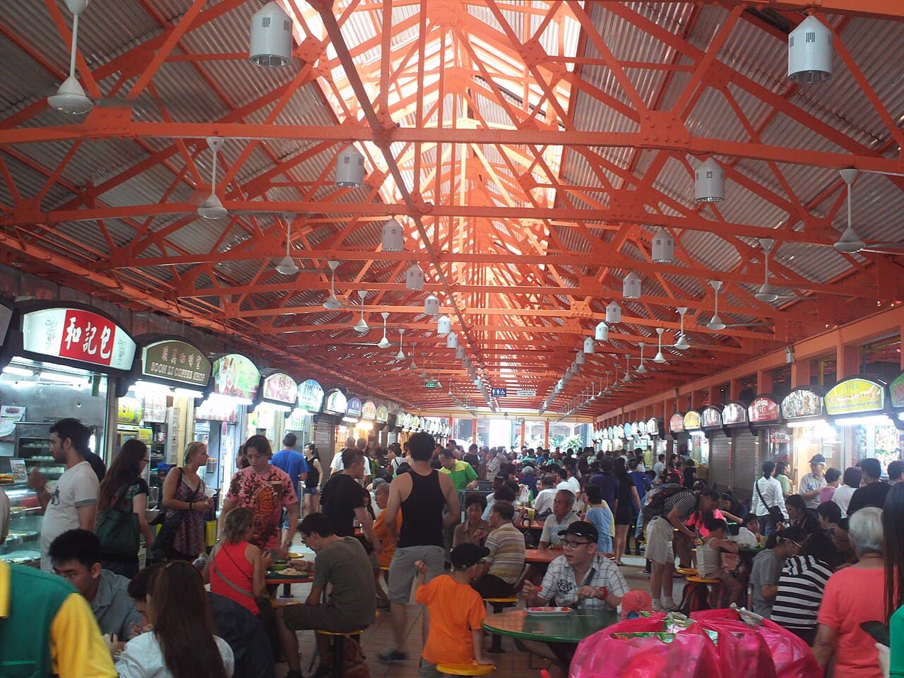 A crowded hawker centre at lunchtime with long queues and diners of all ages sharing tables.
